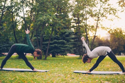 Female yoga student is exercising outdoors with instructor stretching legs and back bending forward standing on mats on green and yellow meadow in city park.