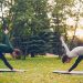 Female yoga student is exercising outdoors with instructor stretching legs and back bending forward standing on mats on green and yellow meadow in city park.
