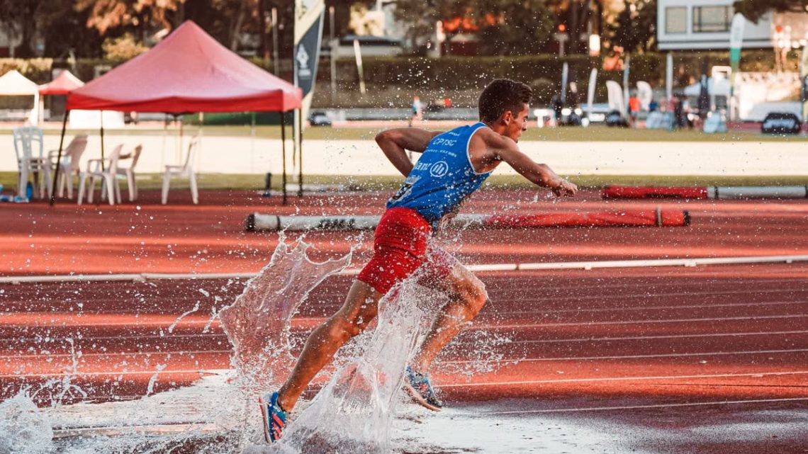Male athlete running through water splash on track field during steeplechase event.