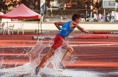 Male athlete running through water splash on track field during steeplechase event.